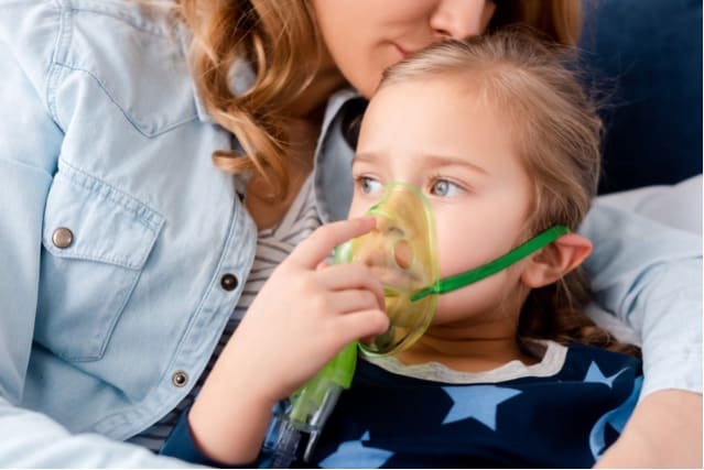 Mother kissing asthmatic daughter in respiratory mask.